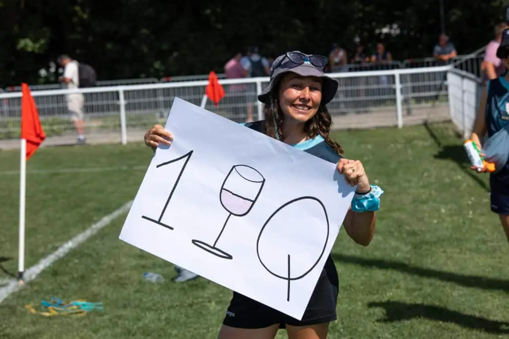 Una aficionada sonriente, con sombrero de sol en la cabeza, levanta al borde del campo un cartel dibujado a mano: un «1», una copa de vino y un balón de rugby