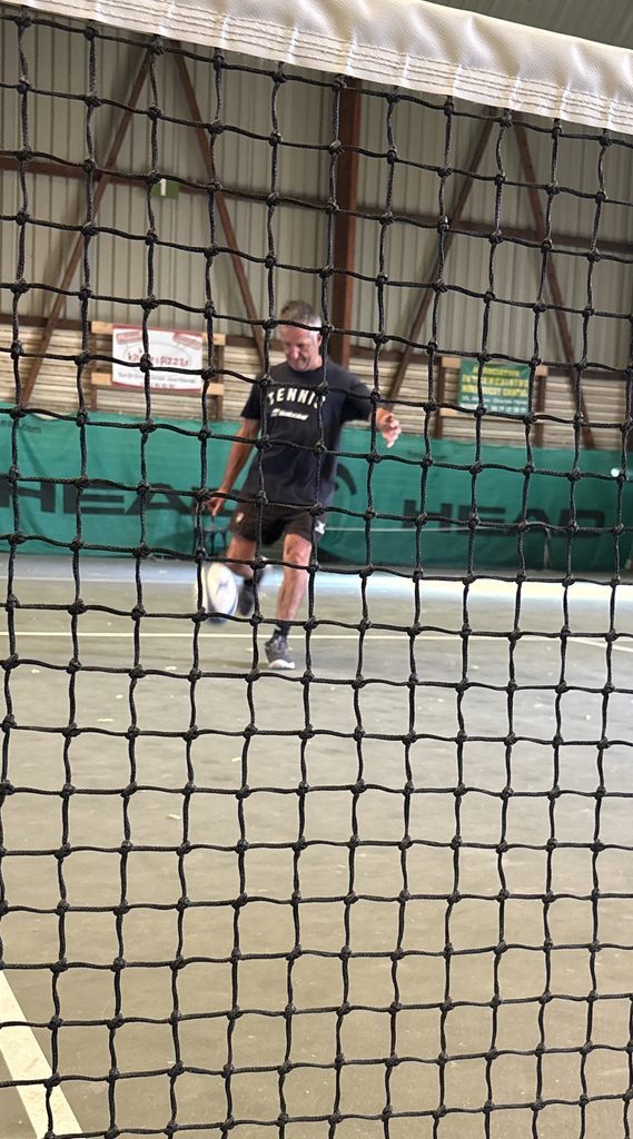 Olivier Miranda golpea con el pie un balón de rugby en una pista de tenis cubierta, visto a través de la red